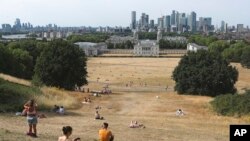 Gente sentada en pasto seco por el sol en el Parque de Greenwich, con el Museo Marítimo y el distrito financiero de Canary Wharf al fondo, en Londres, el domingo 17 de julio de 2022. 