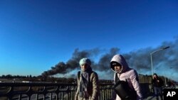 Transeúntes caminan por un puente en Leópolis, en el oeste de Ucrania, mientras una columna de humo se eleva en la distancia tras una explosión cerca del aeropuerto de la ciudad el 18 de marzo de 2022.